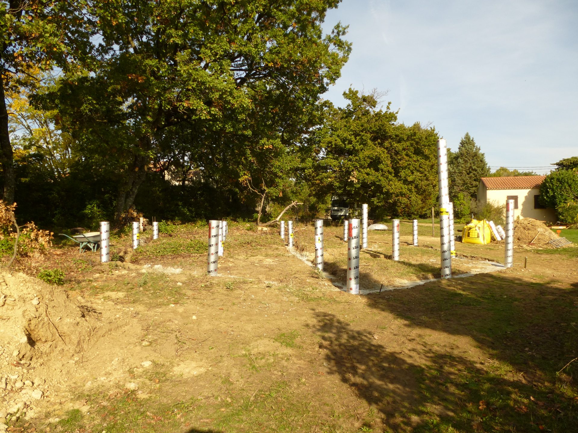 Terrain planté d'arbres où se dresse une trame de tubes-coffrages métalliques verticaux, pour les pieux de fondation.