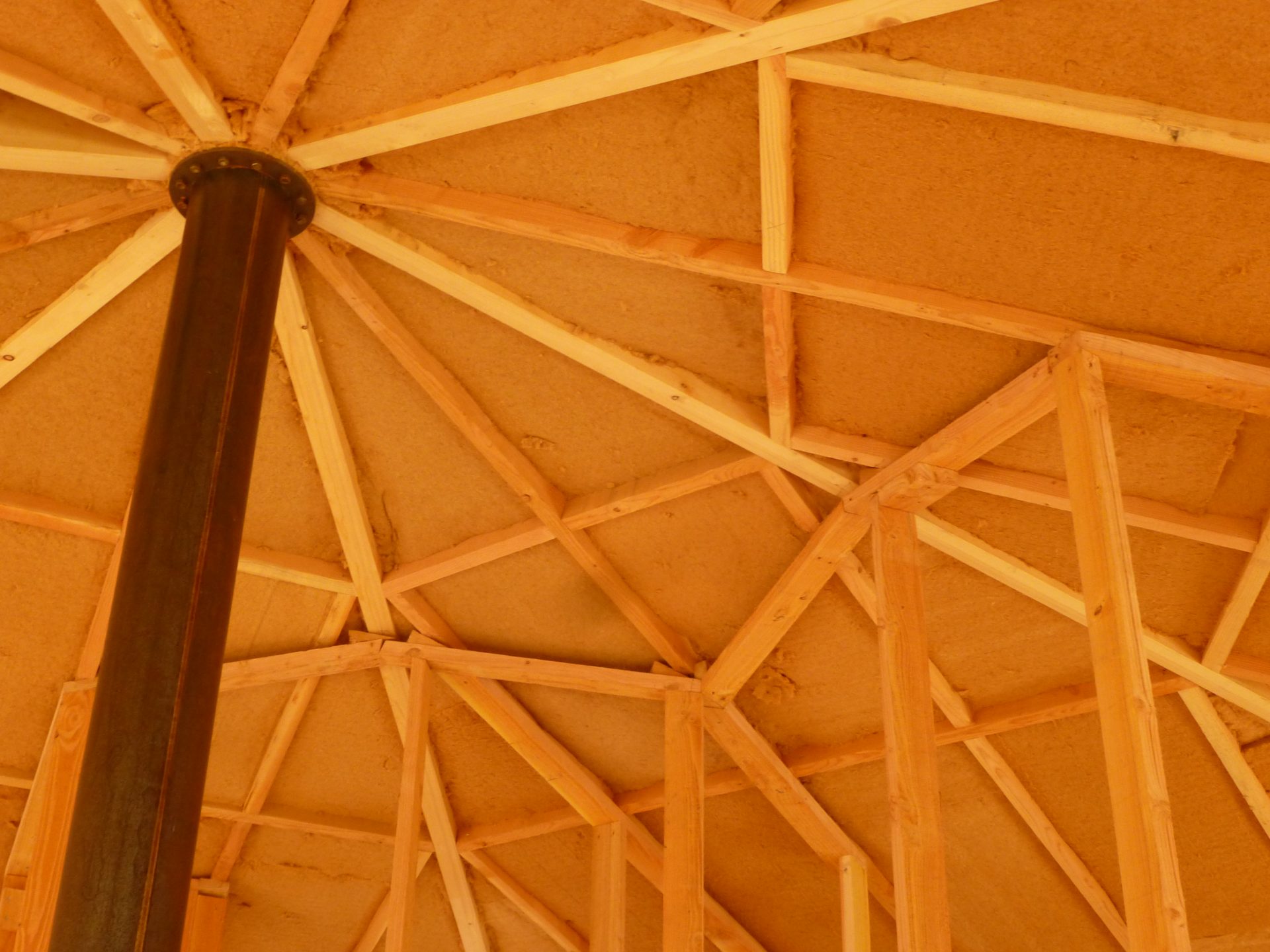 Dessous de la toiture en contre-plongée : chevrons de bois clair rayonnant autour d'un poteau d'acier central, panneaux de fibre de bois.