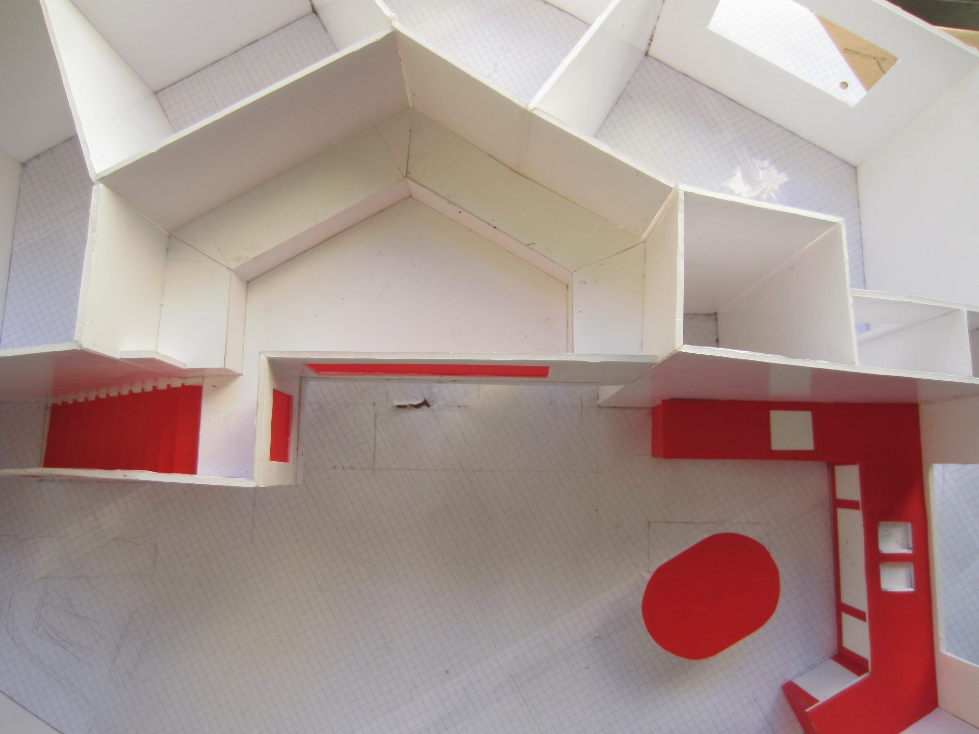 Vue plongeante dans la maquette ouverte : escalier rouge à gauche, bloc cuisine et table ovale rouges à droite, murs blancs.