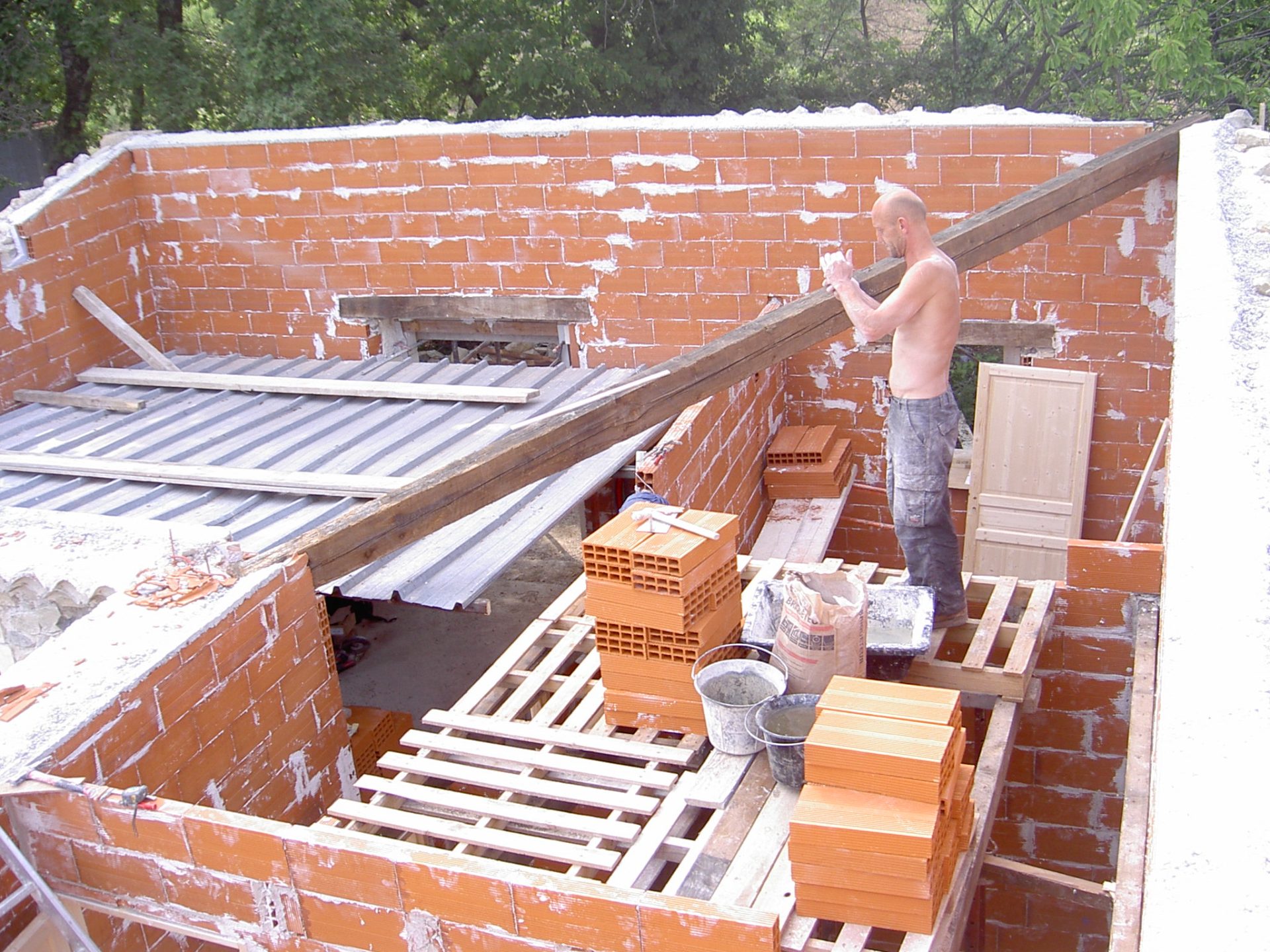 Mazet en construction vu de haut : un maçon travaille en hauteur sur les murs en briques creuses ; palettes de briques, plaques ondulées.